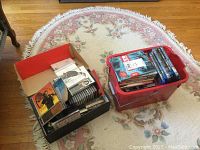 Photo shows two containers on a floral rug: one red plastic bin filled with DVDs and Blu-ray cases and one open cardboard box containing CDs and Nintendo DS cartridges.