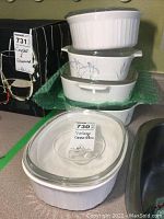 Five stacked vintage Corning Ware casserole dishes with lids. The top three dishes are stacked one inside the other, placed on green bubble wrap next to a black dish. One dish shows floral pattern.