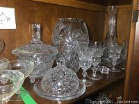 Wide shot of crystal items on wooden shelf including decanter, vase, butter dish, candy dish, and glasses.
