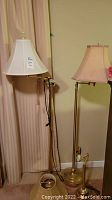 Two brass standing floor lamps with different beige fabric lampshades, one with a pull chain, placed in a corner next to a curtain.