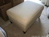 Top and angled front view of rectangular beige upholstered ottoman with dark wooden feet on carpeted floor.