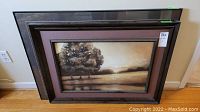 Two framed wall art prints leaning against a wall on a wooden floor. The front print showing a landscape with a large tree and some smaller trees in the background. The frames have dark wood with dual matting in brown and mauve tones.