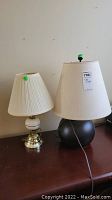 Two table lamps side-by-side on a wooden table, one brass and white ceramic with pleated white shade, the other black base with off-white shade and green knob.