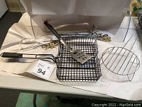 Grilling accessories arranged on white surface including basket, round rack, spatula, and skewers.