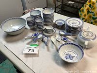 Full view of dinnerware set items laid out on table showing bowls, plates, tea pot, tea cups and spoons with blue floral and geometric patterns on white ceramic.