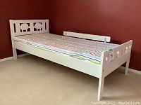 White wooden child's bed with headboard featuring sheep cut-outs, side safety rail, and mattress covered by striped duvet side.