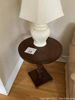 Side view of the dark stained solid wood side table with white lamp on top, showing the table's round top, pedestal base, and square base support.