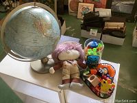 Photo of light blue globe on a silver stand with a stuffed doll with pink hair and beige outfit, and several small toys and containers of craft supplies.