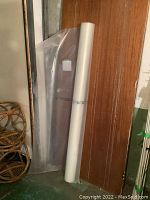 Roll of clear poly tarp leaning against a wooden door with a wicker basket nearby, clear translucent polyethylene sheeting visible attached to roll.