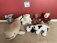 Photo showing all six stuffed animals lined up against a red wall on a carpeted floor: shark, elephant, white bear, small brown bear, white dog with black spots, and plaid-shirt teddy bear.