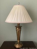Single brass-colored table lamp with detailed ornamental base and off-white pleated shade with pink trim and clear finial on top.