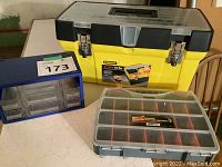 Yellow Stanley plastic toolbox with black and clear lid, blue plastic storage organizer, and gray plastic storage box on white table surface