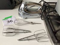 Vintage Oster hand mixer with chrome finish along with two metal beaters and one metal whisk attachment, plated and detachable, placed on a kitchen surface.