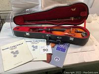 Photo of violin inside hard shell case with bow overhead and three Suzuki Violin School music books laid in front.