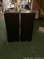 Pair of tall rectangular floor loudspeakers with black fabric grille front, standing upright on the floor.