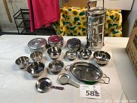 Full view of assorted stainless steel bowls, covered buffet dish, small ladle, and multi-tier tiffin containers along with small trays and lids on a white surface