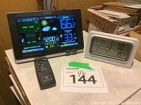 La Crosse Technology wireless forecast station with color display showing weather info, time and date, accompanied by a small black remote control, and an Acurite white digital temperature meter with temperature and weather icons.