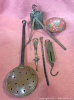 Top-down view of brass and copper items including perforated pan with wooden handle, cow bell, brass measuring dividers, spring scale, and decorative bowl.