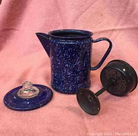 Blue speckled graniteware coffee percolator separated into main pot, lid with glass knob, and metal filter basket parts on pink cloth background.