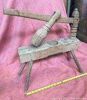 Full side view of antique mechanical vegetable masher showing wooden base, four turned legs, lever arm with pounding head, and vertical screw-like columns.