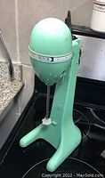 Side view of the mint green Hamilton Beach Drink Master Classic mixer showing egg-shaped head with logo band and attached mixer rods.