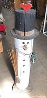 Front view of the handmade log pole snowman showing details of face, buttons, top hat with red bird, and holding bundle of sticks.