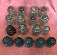 Photo showing 17 telegraph insulators arranged in rows on pink fabric background. Includes a variety of glass insulators in clear, light green, and dark green colors, one smaller mini insulator, and one ceramic insulator.