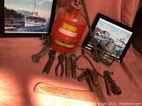 Lot overview showing metal tools arranged in front of two framed boat prints and an orange safety gas can on an orange cloth background