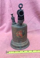 Front view of the brass roofers torch showing cylindrical tank, worn label, valve control, and nozzle opening.