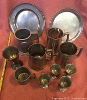 Twelve assorted pewter items including plates, mugs, goblets, and egg cups arranged on a red cloth.