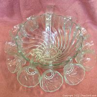 Overhead view of clear glass punch bowl with swirl pattern and twelve matching cups arranged around it on a pink surface