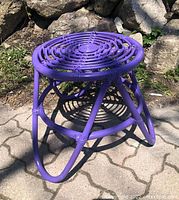 Front angled view of purple rattan side table showing woven top and curved tubular legs with shadow on ground