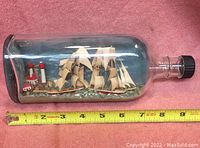 Side view of the glass bottle clearly showing the ship with sails, lighthouse, and house inside, alongside measurement tape showing bottle length around 9 inches.