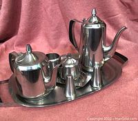 Photo showing entire stainless steel tea and coffee set on oval tray, including teapot, coffee pot (lid detached), creamer, sugar bowl with lid, salt and pepper shakers.