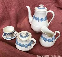 Full view of the five-piece Wedgwood Queensware tea service showing the teapot, creamer, sugar bowl with lid, tea cup, and saucer with blue embossed garland pattern.