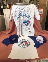 Four-piece Toronto Blue Jays lot with 2 T-shirts and 2 hats displayed. Signed jersey T-shirt with Blue Jays logo, Home Opener 92 T-shirt, and two hats with Blue Jays logos.