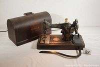 Singer antique sewing machine with attached curved metal motor starter on wooden base adorned with gold scrolls and florals, positioned next to bentwood case with handle.