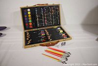 Opened wooden art box displaying oil pastels, watercolor paint palette with 12 colors, various colored pencils organized in rows, and small paint brushes placed outside the box.