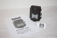 Bushnell X-8 Trail Cam with instruction manual and lot number sign in background.