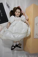 Porcelain doll in a white dress with black shoes, original box standing beside it, certificate and tags visible nearby.