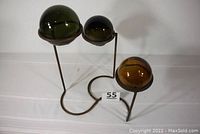 Three glass witch balls (green, black, amber) displayed on a dark iron stand. The stand has three prongs at different heights to hold each ball.