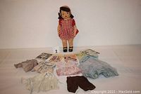 Photo of the Terri Lee doll standing wearing red and white gingham dress with multiple extra outfits laid out in front of it on a flat surface.