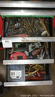 Three metal drawers containing various tools including drill bits, clamps, pliers, and other hand tools with signs of rust and wear.