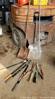 Multiple garden tools laid out on dirt floor in front of a forklift, showing shovels, rakes, Fiskars pruners, and hand tools.