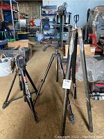 View of all three camera stands, showing one wooden stand with chain and damage, and two metal tripods including an Opitex T300 tripod and another Manfrotto tripod.