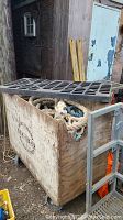 Wooden crate with removable slotted lid partially open showing coiled ropes inside, placed outdoors with surrounding items.
