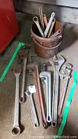 Photo of various metal wrenches arranged on floor and inside a bucket, showing multiple sizes and types including pipe wrenches and combination wrenches.