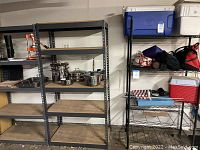 Photo showing two large metal racks with pressboard shelves holding kitchenware and a black wire rack shelf on the right with coolers and various items.