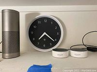 Photo of Amazon Echo Wall Clock with black face and white frame, next to 3 Amazon Echo units on a shelf, showing overall parts of the lot