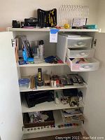 White cabinet open showing shelves with batteries, stationery supplies, tool kit, magnifying glasses, and office accessories.